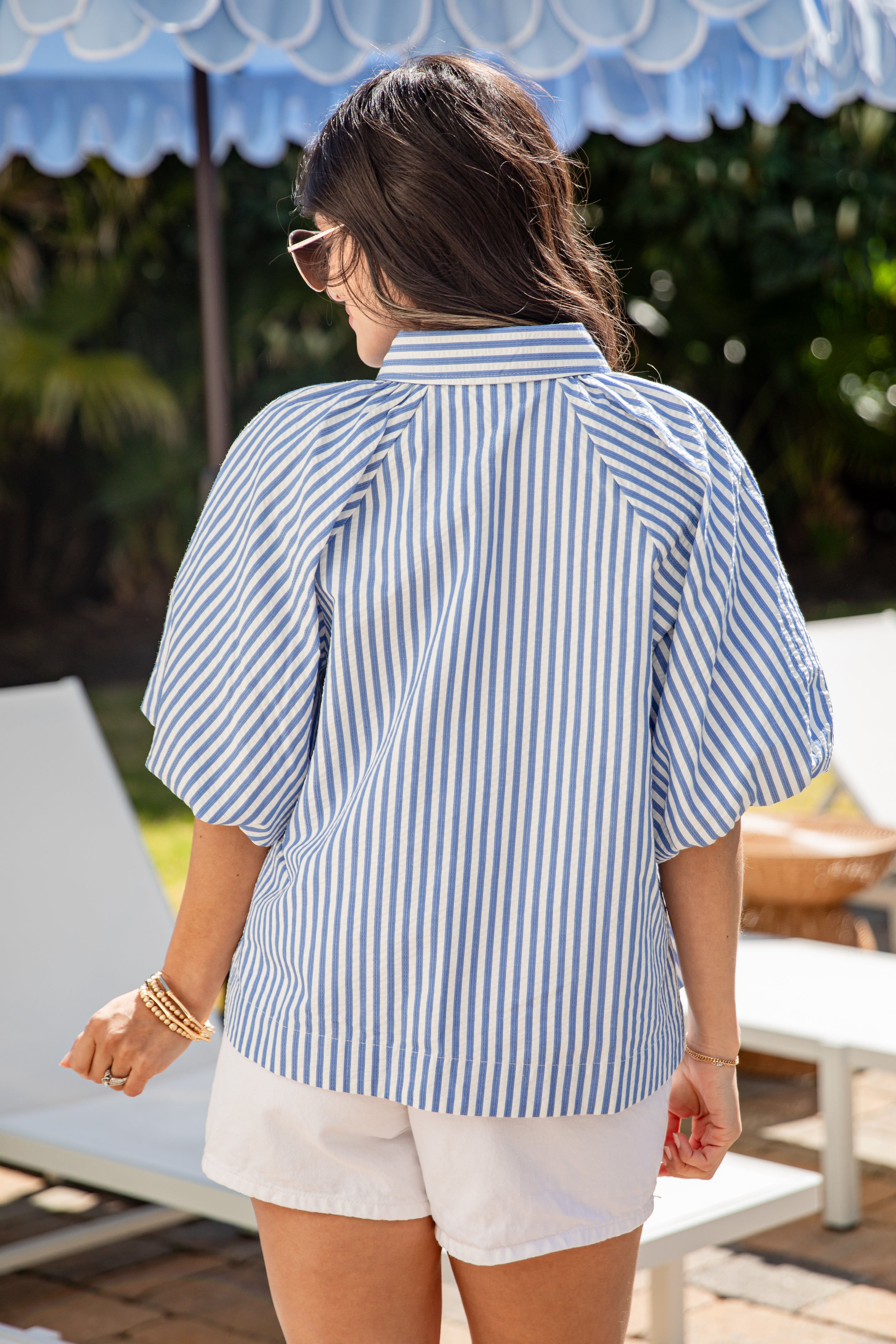 Woman wearing a blue and white striped shirt and white shorts, standing under a blue umbrella.
