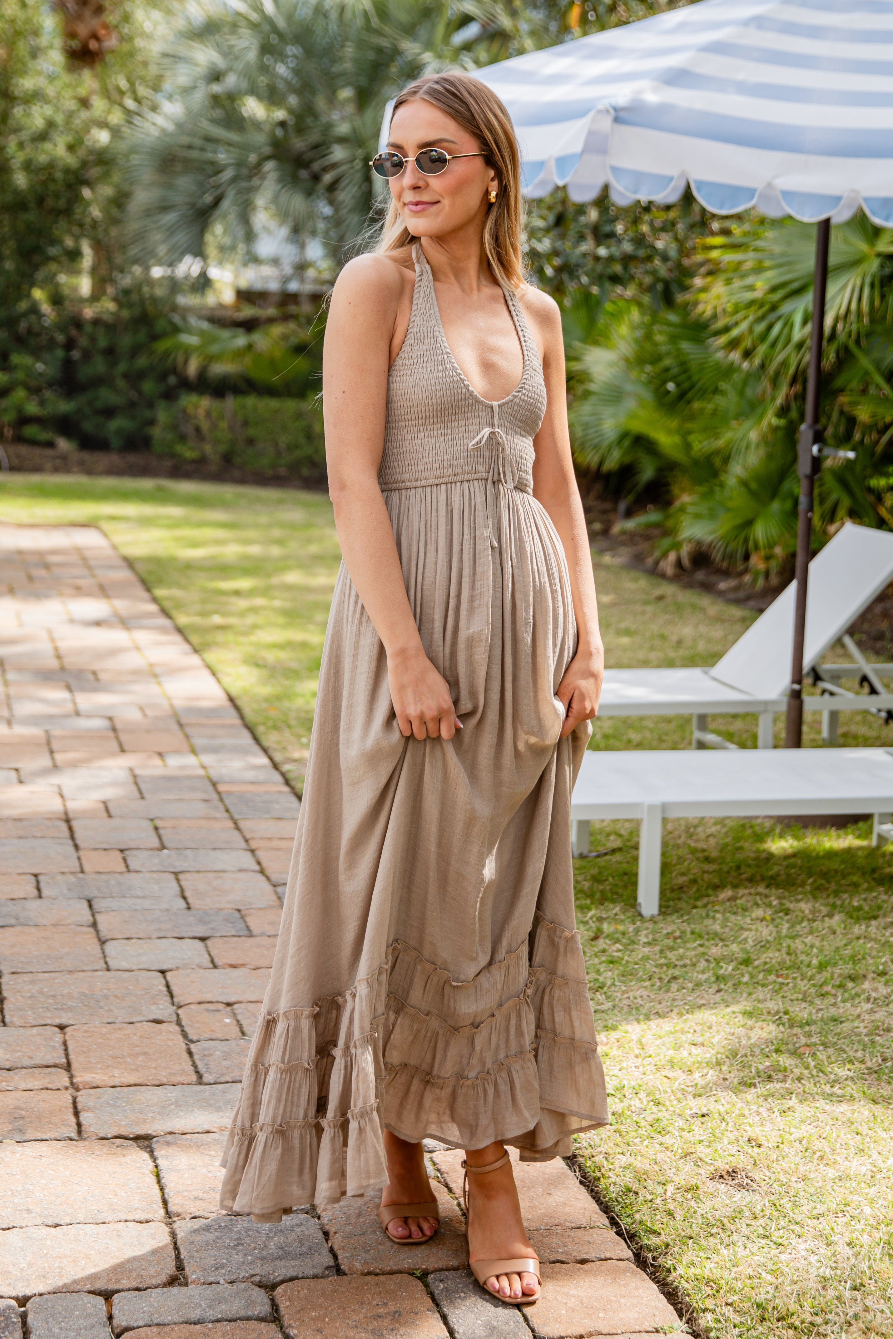 Woman in a light dress standing under a striped umbrella in a garden setting.