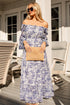 Woman in a blue floral dress holding a beige clutch on a wooden deck.