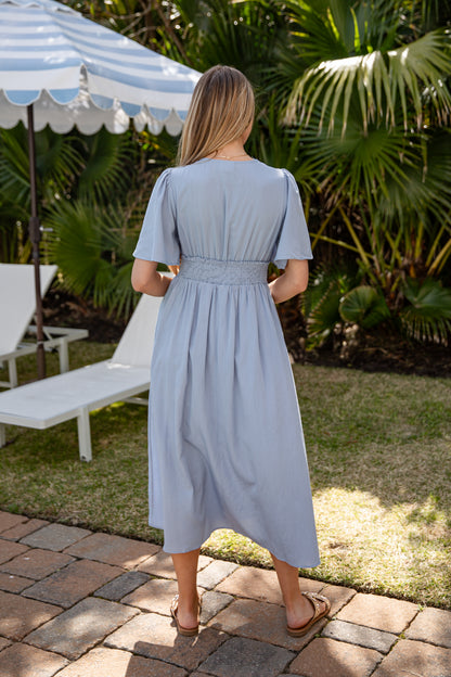 Woman in a light blue dress standing on a patio with greenery and a striped umbrella in the background.