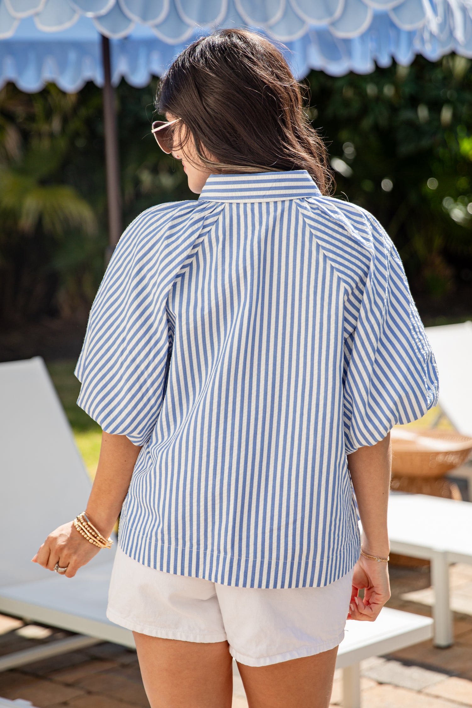 Woman wearing a blue and white striped shirt and white shorts, standing under a blue umbrella.