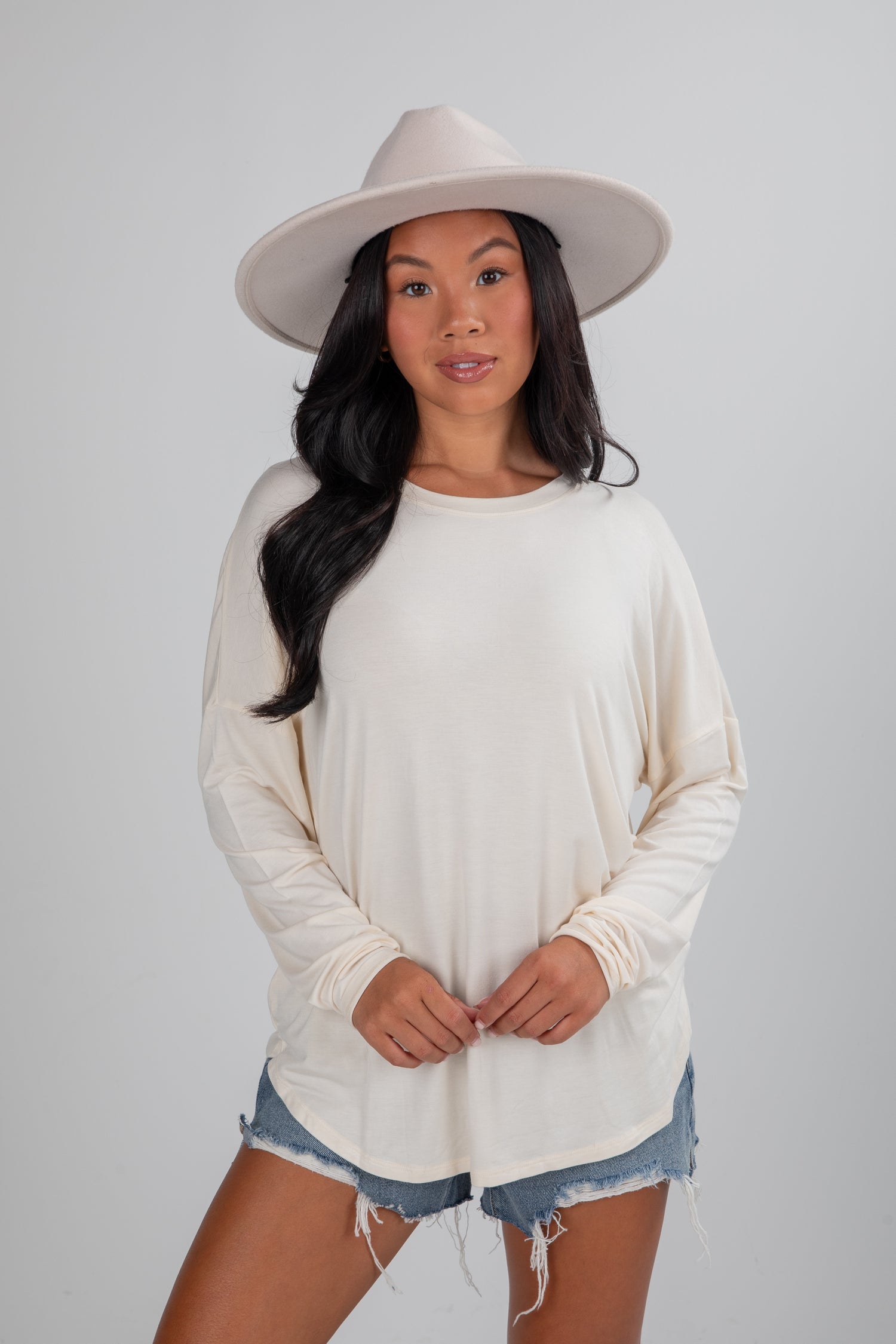 Woman wearing a beige hat, white long-sleeve top, and denim shorts on a plain background