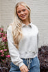 Woman wearing a light gray cropped hoodie and blue jeans standing in front of a white wall with flowers.