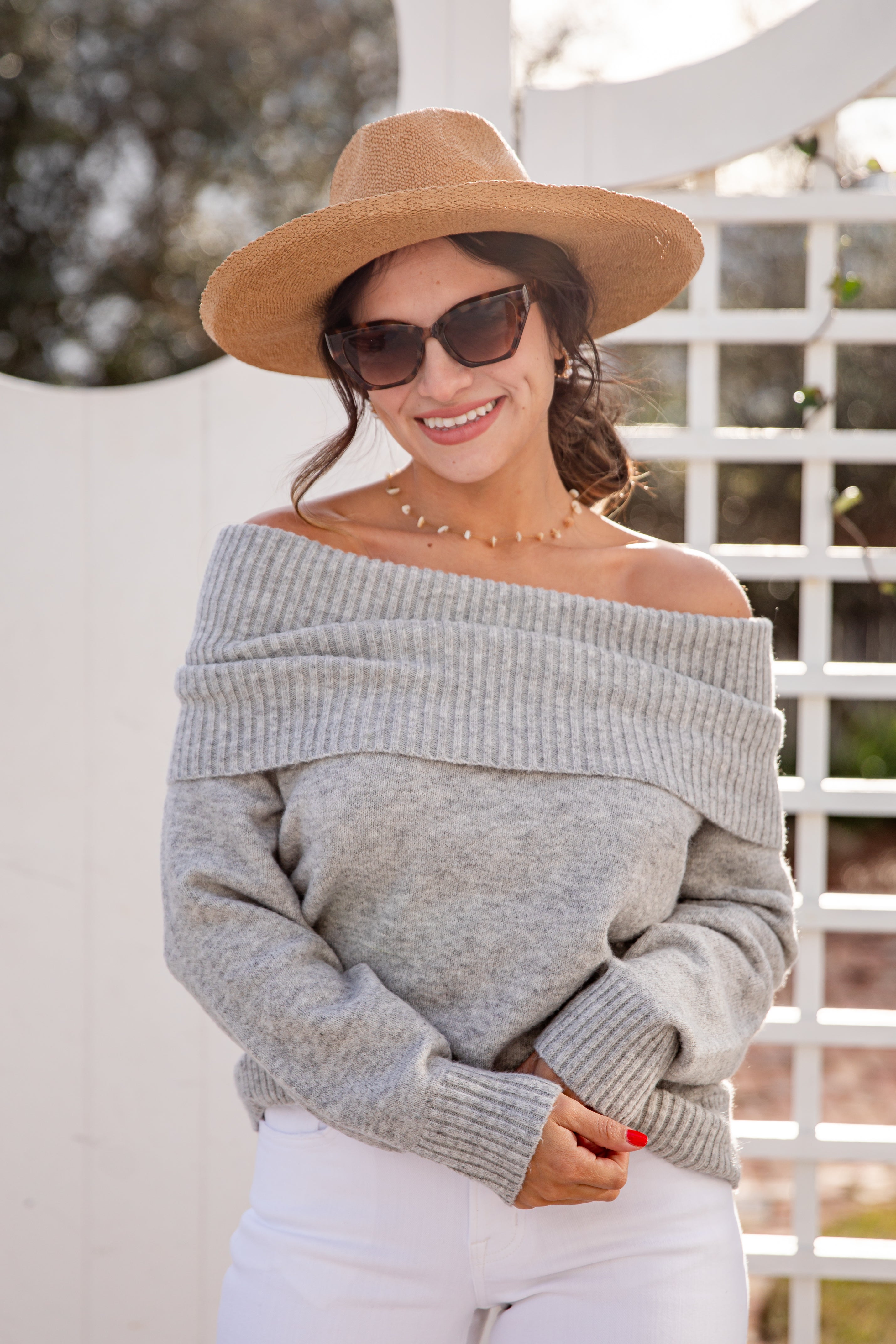 Woman wearing a gray off-shoulder sweater, white pants, and a brown hat with sunglasses.