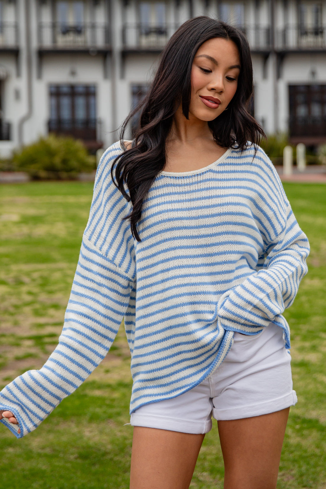 Woman wearing a blue and white striped sweater and white shorts standing in a grassy area with a building in the background.