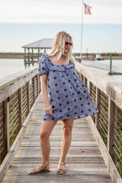 Woman in a blue floral dress standing on a wooden dock by a body of water.