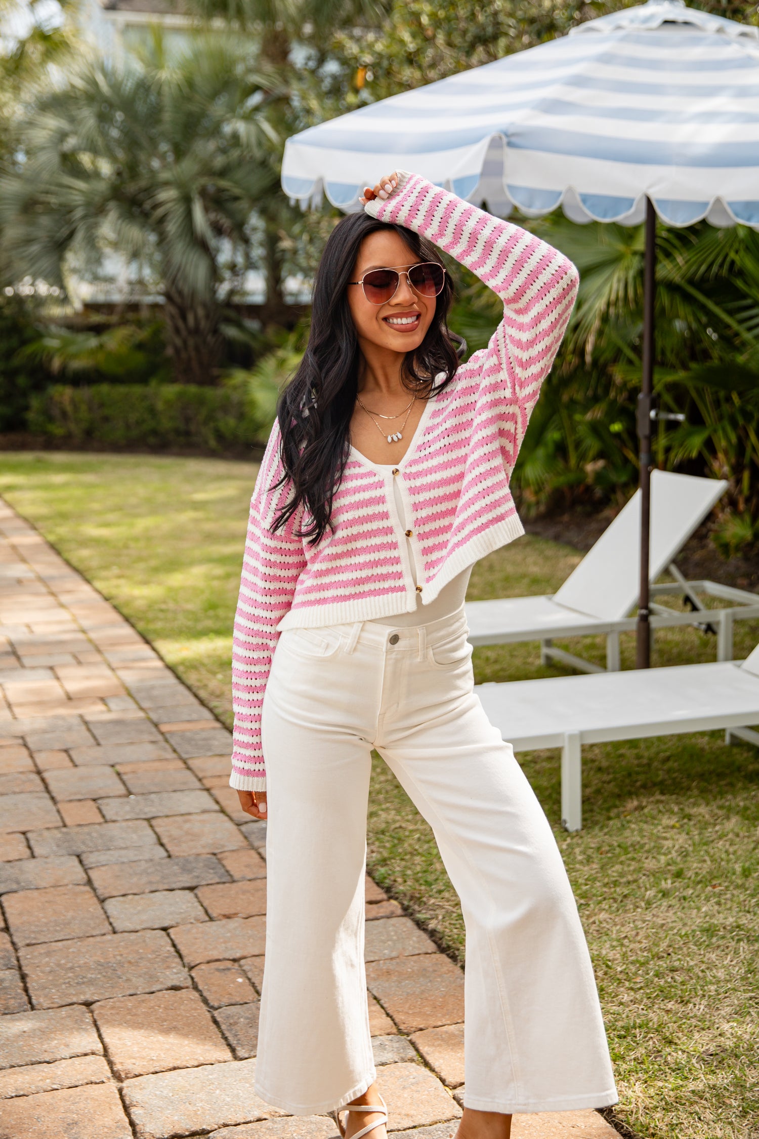 Woman in a pink and white striped cardigan and white pants standing outdoors with a striped umbrella in the background.