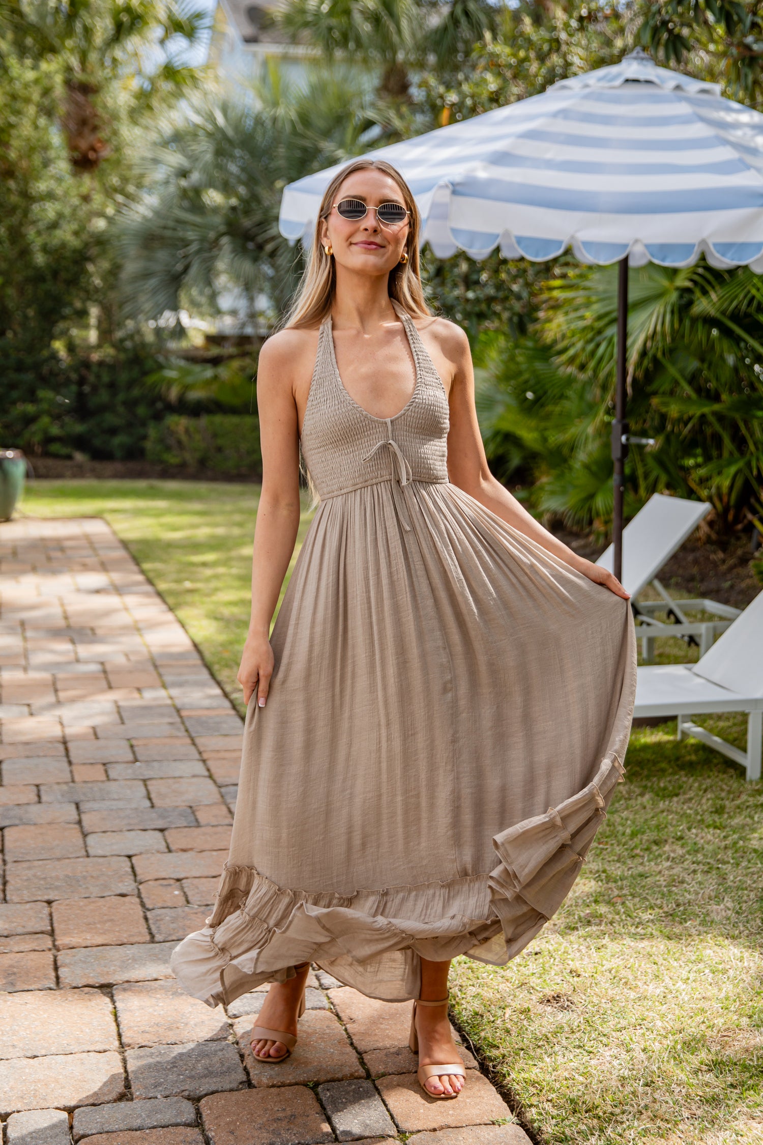 Woman in a light dress standing under a striped umbrella in a garden setting.