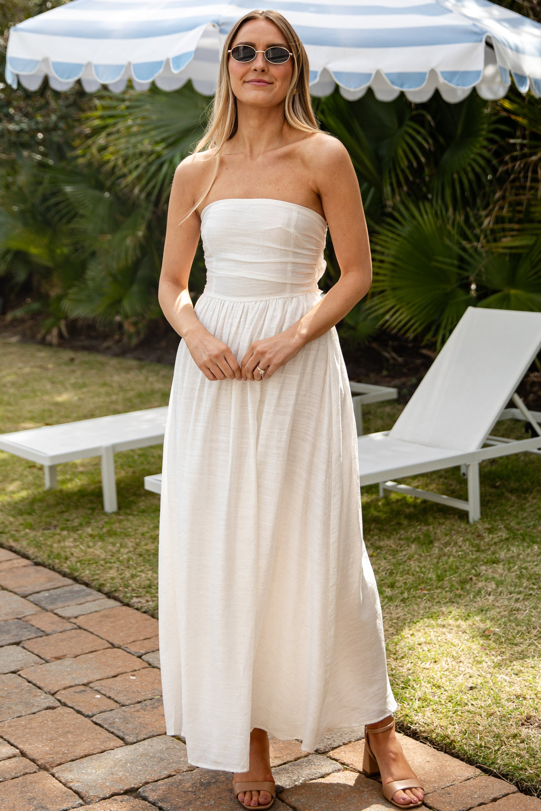 Woman in a strapless white dress standing under a striped umbrella in a garden setting.