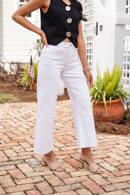 Person wearing a black top with gold buttons and white pants standing on a brick patio.