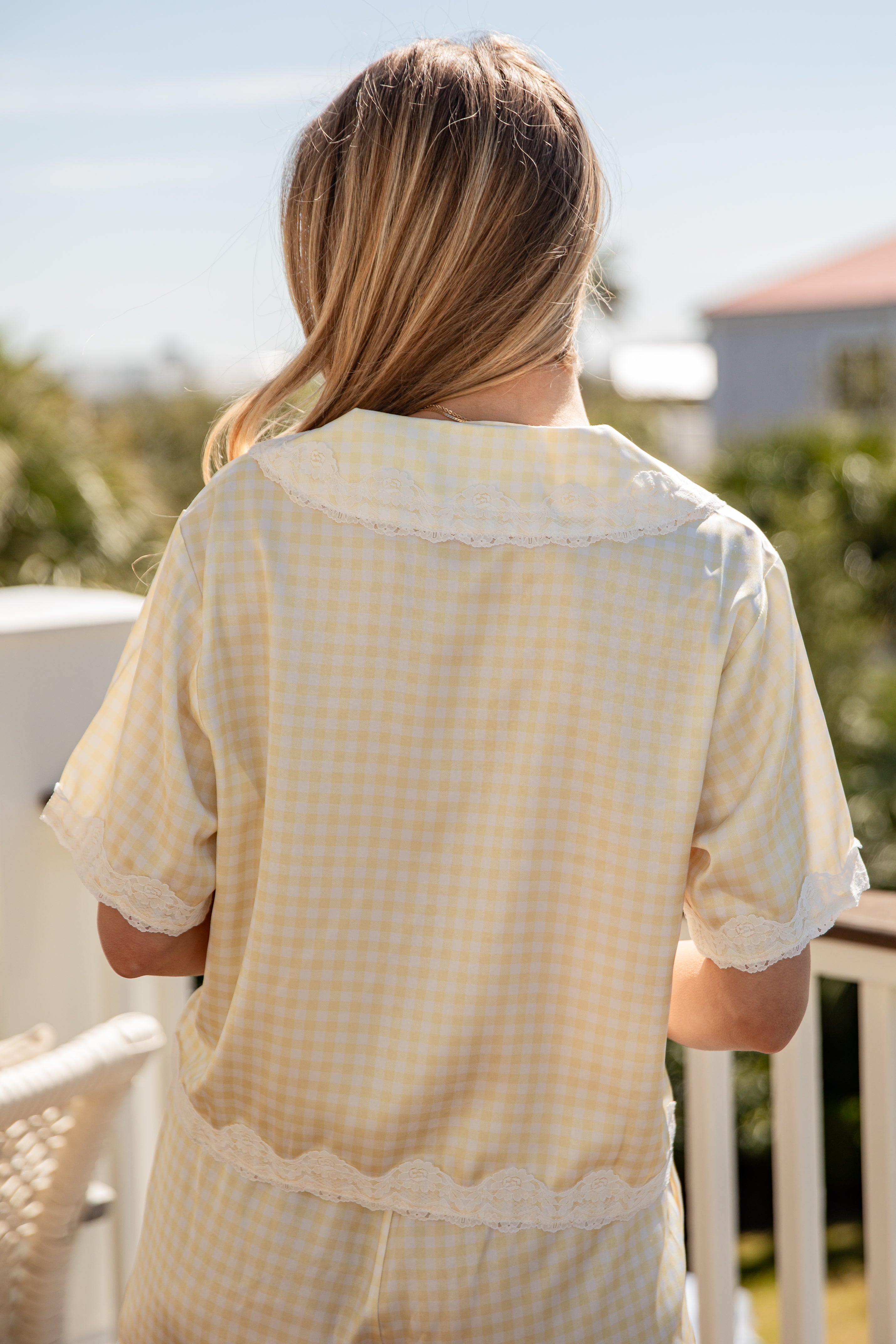 Woman wearing a light yellow checkered shirt on a balcony with greenery and a clear sky.