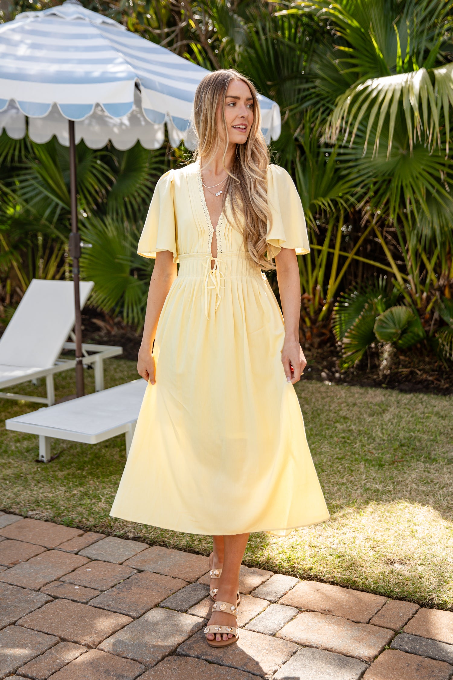 Woman in a yellow dress standing outdoors with greenery and a striped umbrella in the background