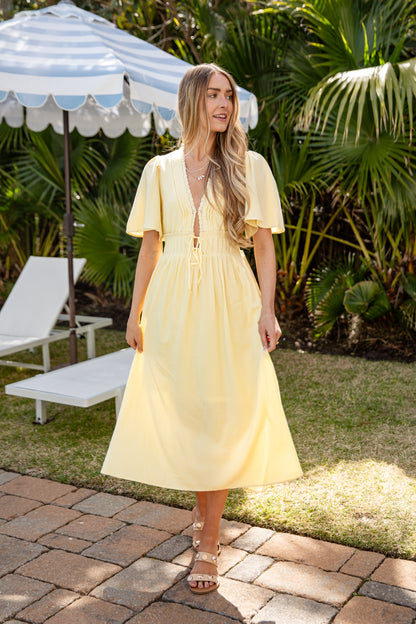 Woman in a yellow dress standing outdoors with greenery and a striped umbrella in the background