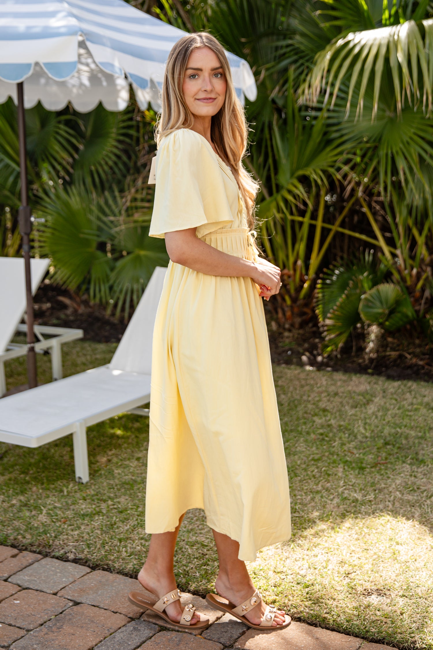 Woman in a yellow dress standing outdoors with greenery and a bench in the background
