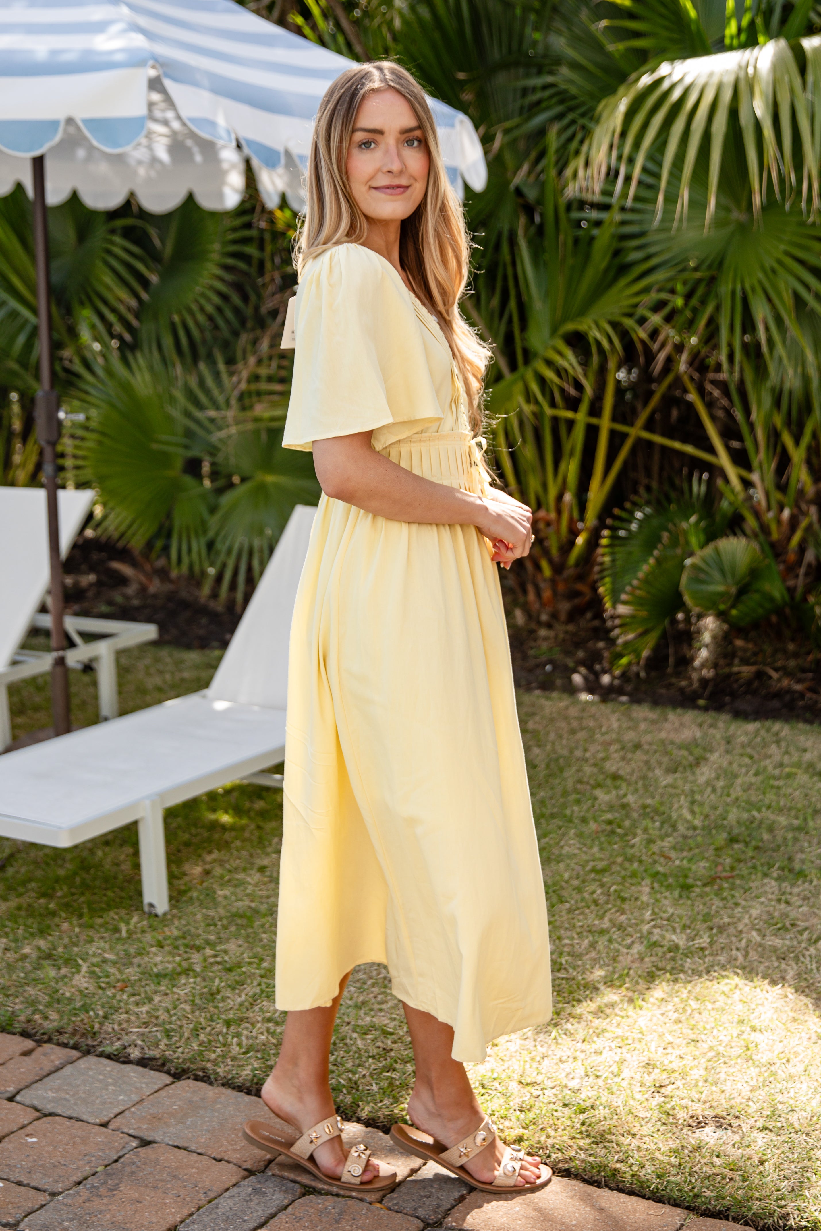 Woman in a yellow dress standing outdoors with greenery and a bench in the background