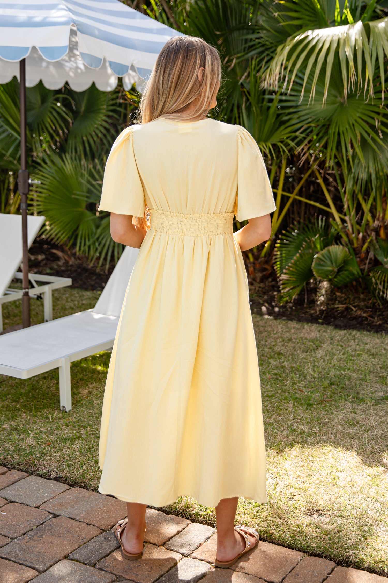 Woman in a yellow dress standing outdoors with palm trees and a white umbrella in the background.