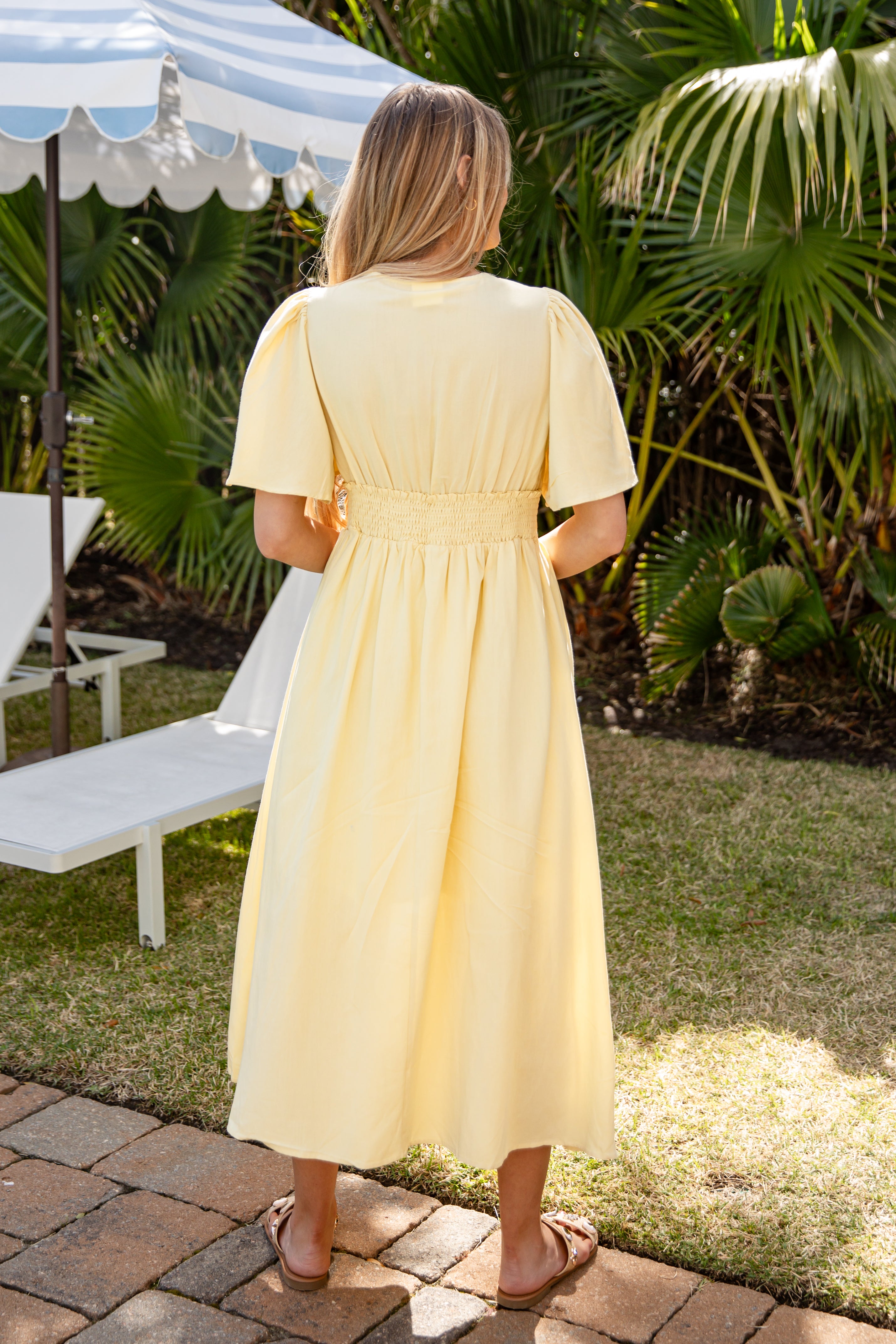 Woman in a yellow dress standing outdoors with palm trees and a white umbrella in the background.
